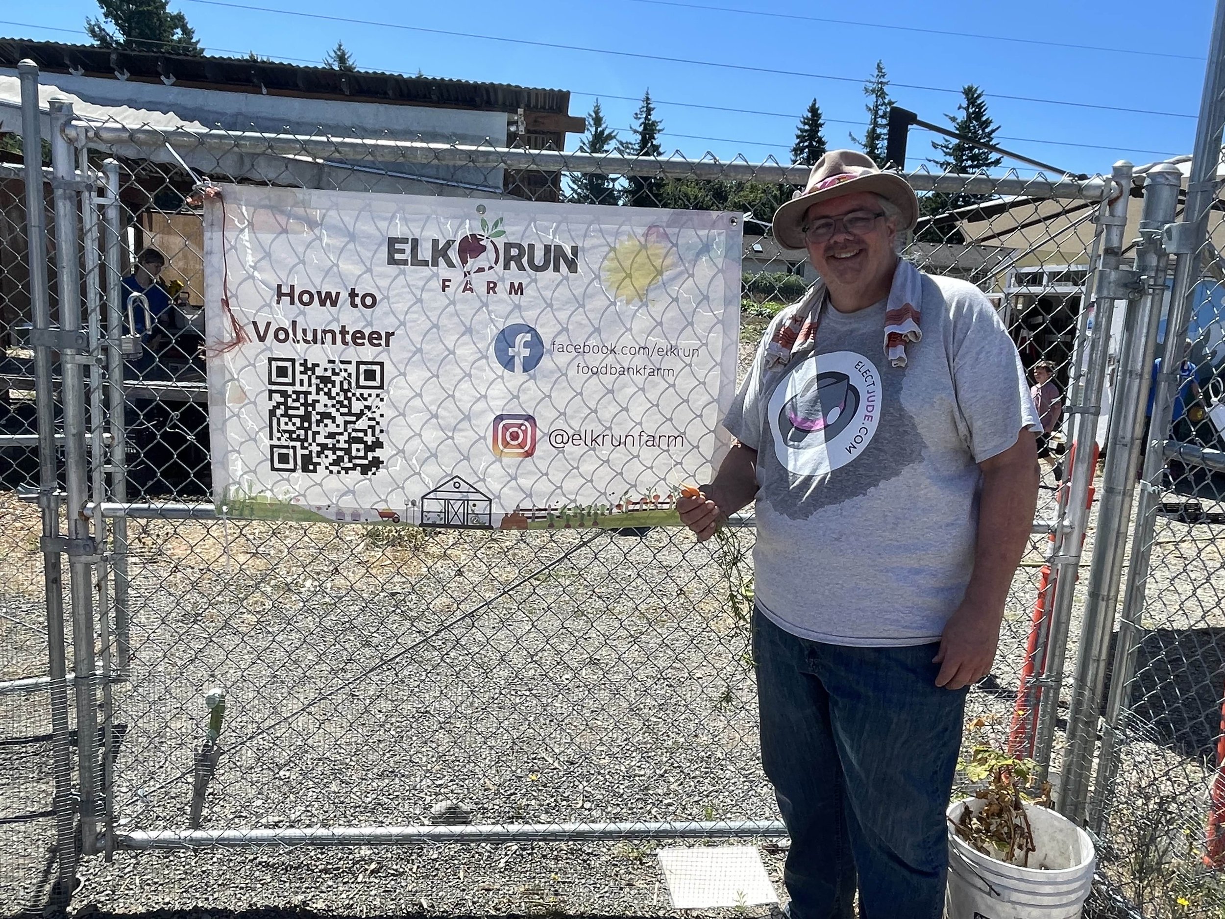 Jude Anthony, in iconic hat and tee, soaked from effort and cooling water, holding treats for his pets in front of the Elk Run Farm sign.