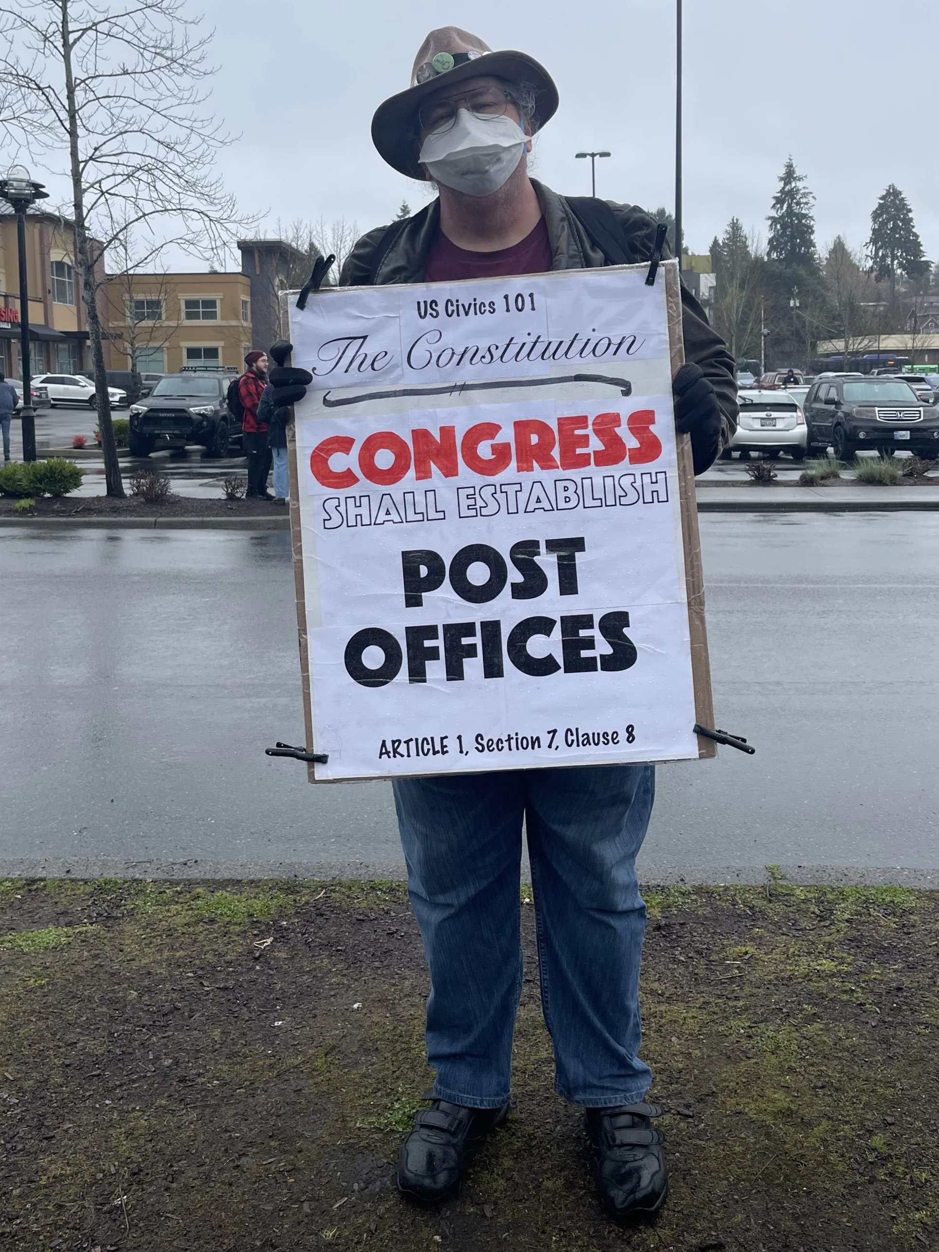 Jude Anthony, a tall white man wearing KN95 mask, glasses, and his iconic hat holding a homemade posterboard protest sign that says "US Civics 101 The Constitution. Congress shall estabilsh post offices. Article 1, Section 7, Clause 8"